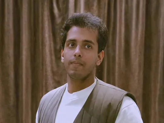 Dev, a young man with short dark hair, in a white top and a brown quilted vest, in front of a brown curtain.